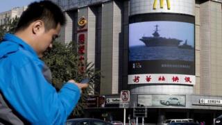 A Chinese man checks his mobile phone near a huge screen showing a news report of the deployment of US aircraft carrier USS George Washington to typhoon-hit Philippines on 14 November 2014