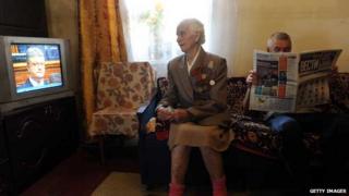 Elderly woman watches TV while man reads a newspaper