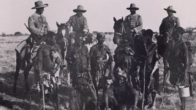 Hombres Herero a pie demacrados y encadenados al frente de una fila de soldados alemanes a caballo