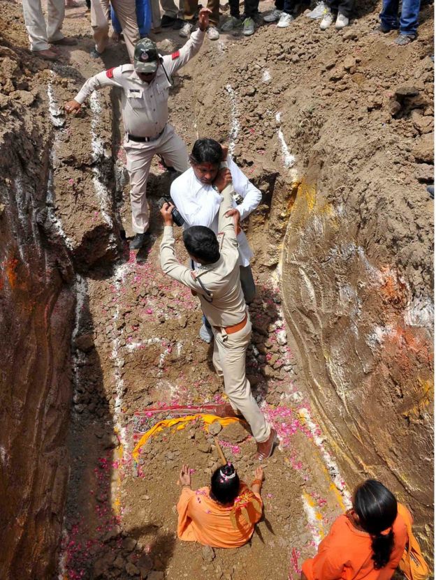 A devotee of Indian Hindu Akhara leader Trikal Bhawanta grapples with two police officials in a deep grave, alongside the seated Trikal Bhawanta, as she undergoes a 