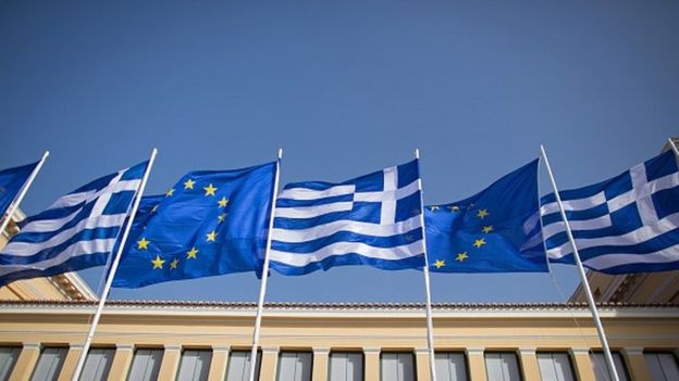 The national flag of Greece and the flag of the European Union fly above a government building.
