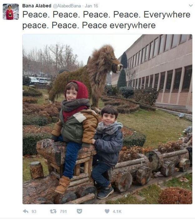 A screenshot from the Twitter account @AlabedBana, showing her two younger brothers smiling and seated on a a row of plant pots made in the shapes of a train. The caption reads 