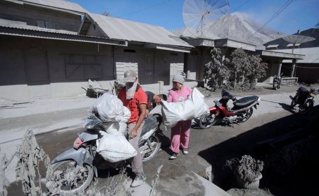 A local couple load their belongings onto motorbikes, surrounded by ash-covered homes, as they evacuate from Gamber Village on 23 May
