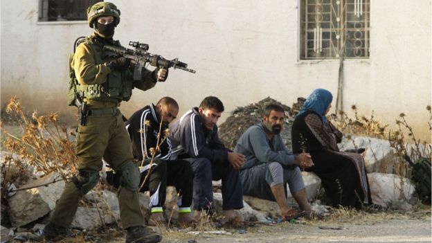 An Israeli soldier keeps watch as Palestinians sit nearby after the army entered the village of Yatta in the occupied West Bank