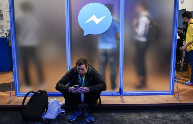 Un hombre con su celular sentado bajo el logo de Facebook Messenger.