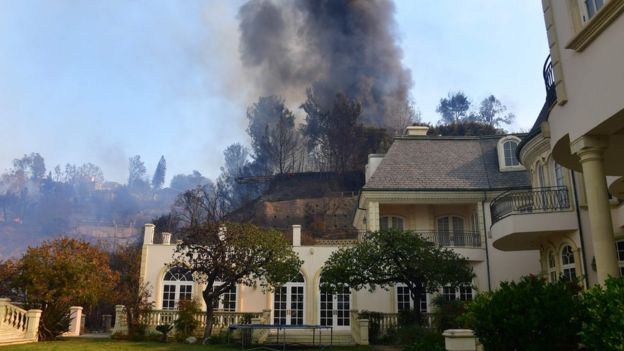 A huge plume of black smoke rises from a burning home on a hilltop beside one still standing in Bel Air, east of the 405 freeway on December 6, 2017 in Los Angeles, California