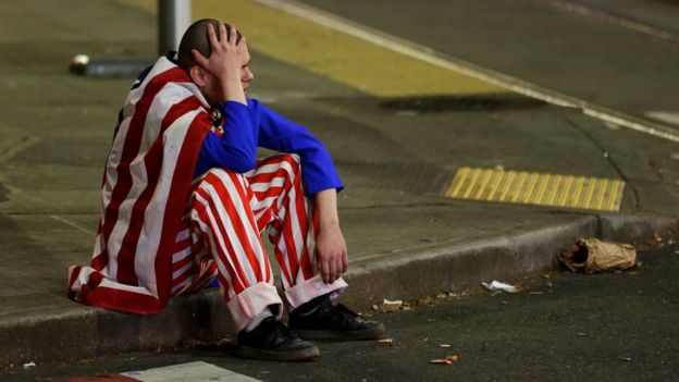 A man dressed in red-white-and-blue sits on the curb during a protest against President-elect Donald Trump