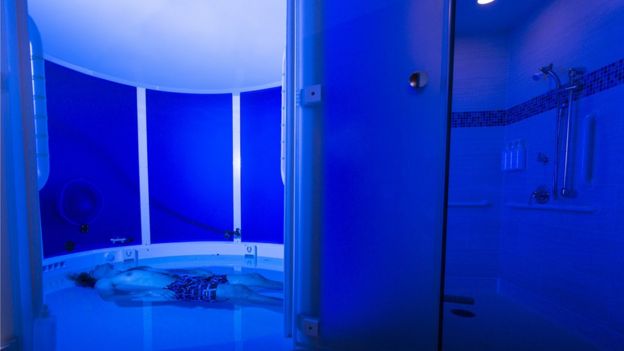 Blue-tinged wide shot of man lying down in large flotation tank