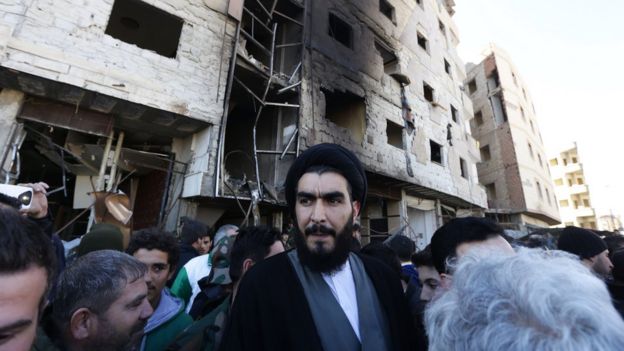 A Shia cleric stands amid Syrian pro-government forces and residents at the site of suicide bombings in the area of a revered Shia shrine in the town of Sayyida Zeinab, on the outskirts of the capital Damascus, on Sunday