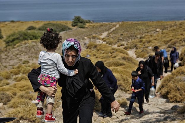 Line of refugee women, some holding children, ascend hill from beach to road