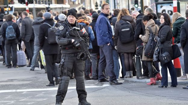 An armed police officer oversees queues of people waiting for a bus
