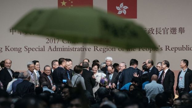 Protesters hold a yellow umbrella and a banner as Carrie Lam, Hong Kong's chief executive-elect, center, stands on stage with her family and supporters after announcement of the chief executive election on March 26, 2017 in Hong Kong, Hong Kong.