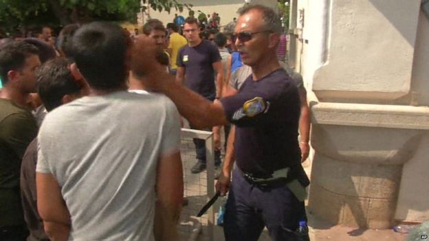 Greek police officer confronting migrants, slapping one while brandishing a knife