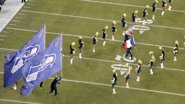 NFL players run onto the field with American and French flags surrounded by cheerleaders