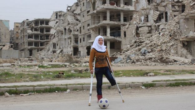 Image of Saja, a Syrian child missing a leg, playing football in front of bombed buildings
