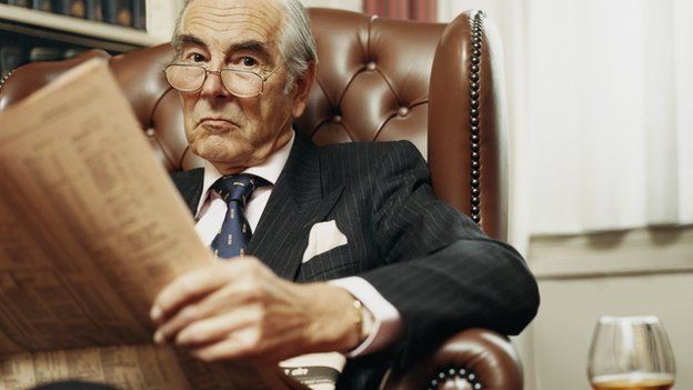 An old man in a suit looks up from his newspaper and brandy.