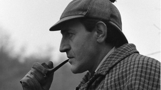 un fotogramma del celebre Sherlock Holmes interpretato da Douglas Wilmer, di profilo fuma la pipa e indossa il classico cappello
