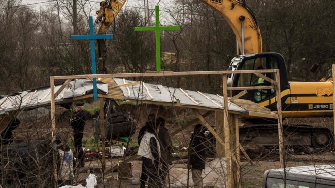 French police officers stand guard as an excavator knocks down buildings including a makeshift mosque and Evangelical Church