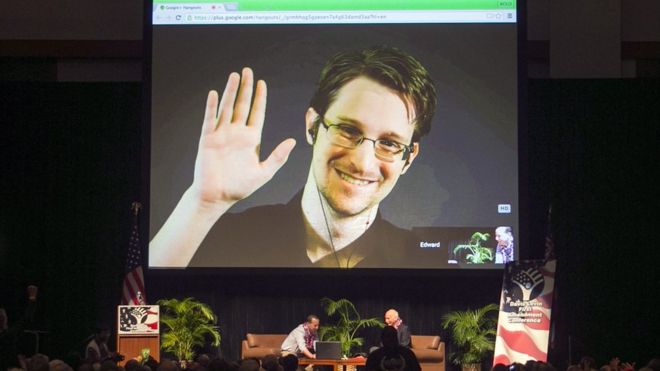 Edward Snowden smiles and waves from a huge screen set up in a conference hall full of people