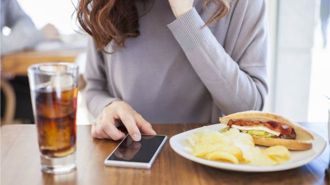 A woman drinking and eating while using mobile phone