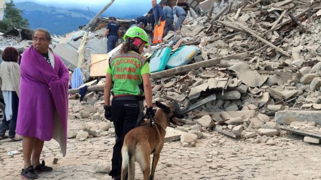 A man wrapped in blanket looks on as a rescuer with a dog stand in front a collapsed house