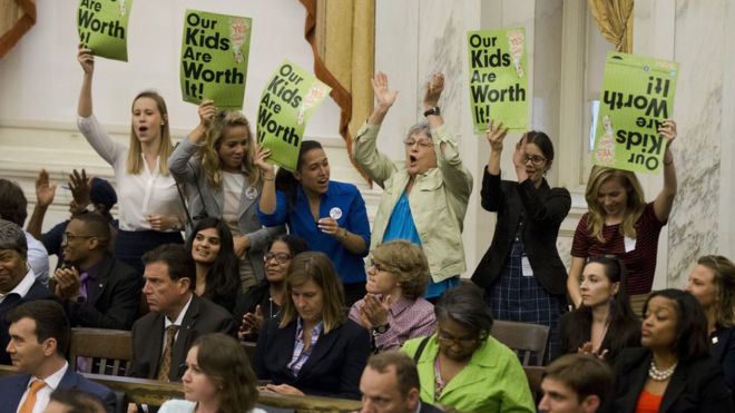 Six woman standing at the back of the chamber cheer, holding signs that say "our kids are worth it"