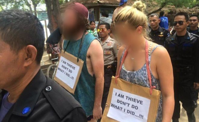 Western man and woman being paraded by Indonesian police and security guards with a sign around their necks reading "I am thieve [sic] don't do what I did...!!!"