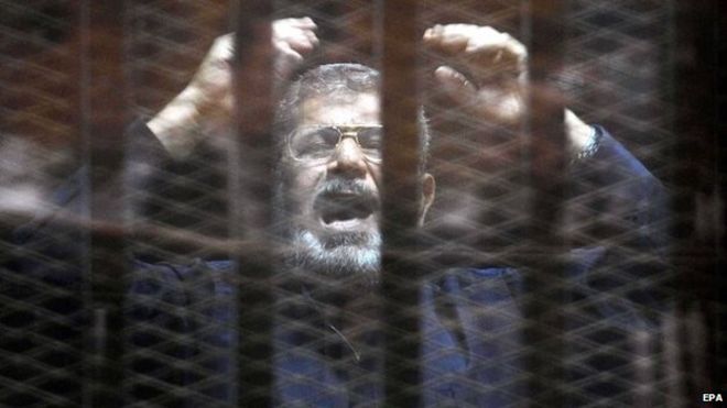 Mohammed Morsi stands inside a glassed-in defendant's cage (16 June 2015)