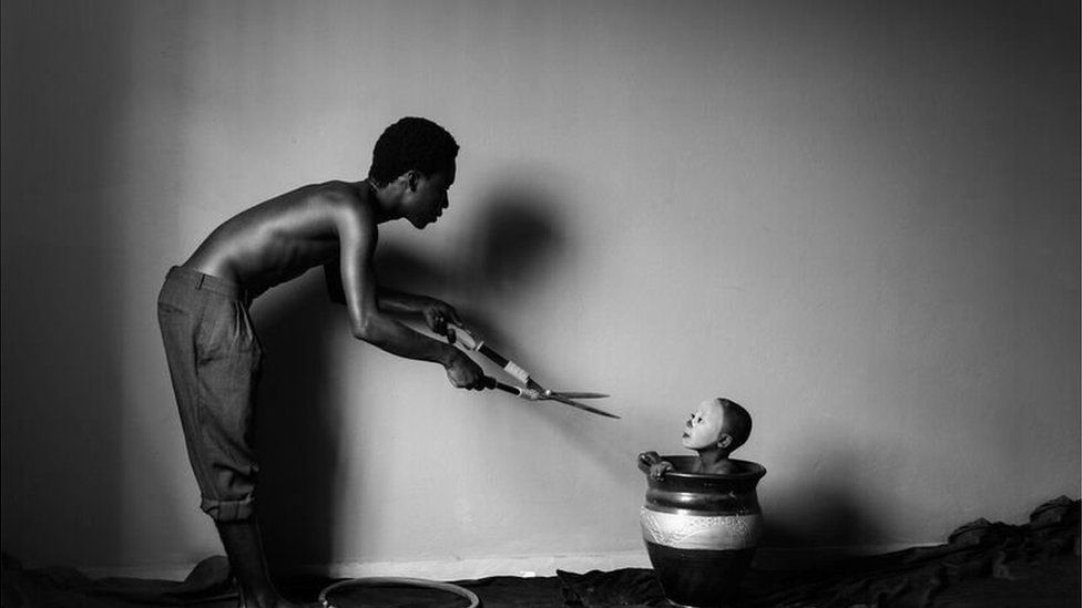 A photo from a series entitled Asylum by Eric Gyamfi from Ghana showing a man with sheers approaching a young boy in a pot