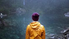 A woman wearing a yellow raincoat and a beanie stands in front of the tarn
