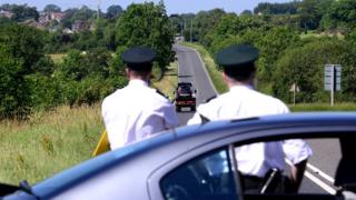 a car is shown being taken away with police officers out of focus looking on