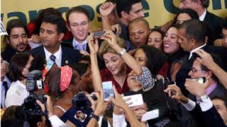 Dilma Rousseff surrounded by a tight crowd of people smiling and taking photos on their smartphones
