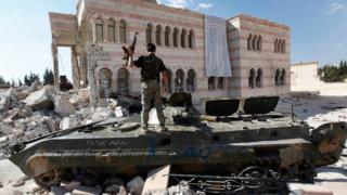 A Free Syrian Army soldier stands on a damaged Syrian military tank in front of a damaged mosque, which were destroyed during fighting with government forces