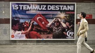 seorang pria berjalan melewati billboard dengan gambar tentara, bendera Turki, dan tulisan 15 Temmuz Destani