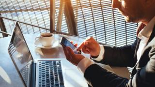 شخص يمسك بهاتفه المحمول الذي يعمل باللمس بينما يجلس أمام جهاز الكمبيوتر المحمول