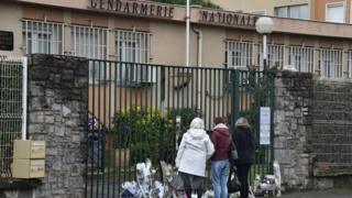 Three women stand outside the Gendarmerie headquarters in Carcassonne, where flowers have been placed