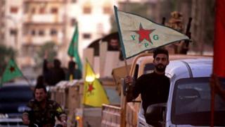 Hombre sosteniendo una bandera con las iniciales de las YPG.