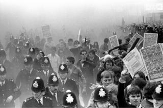 Police protect defiant National Front supporters. Some carry printed placards, “National Front is a Racialist Front Join the National Front.”