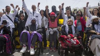 A crowd of people holding their fists high, with the hunger strikers ahead of them in wheelchairs