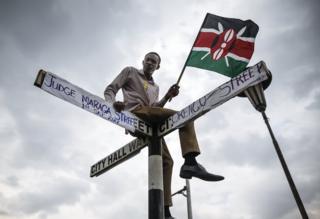 Siku hiyo hiyo, Mkenya anaketi kwenye bango nje ya Mahakama Kuu huko Nairobi ambapo Jaji Mkuu David Maraga alitangaza kufutiliwa mbali kwa matokeo ya kura ya urais katika uchaguzi mkuu wa Agosti . Majina ya barabara kwenye bango yamebadilishwa na watu wanaosherehekea uamuzi huo na kubandikwa ya mwanasheria James Orengo, ambaye alikuwa wakili wa upinzani na Jaji mkuu David Maraga.