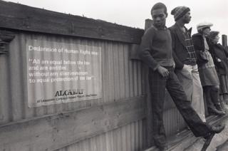 A young man stands next to an ALCARAF sign