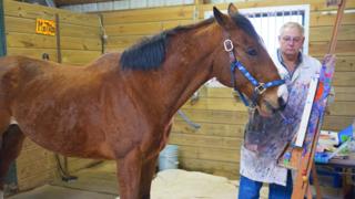 Metro the horse holding a paintbrush in front on an easel, standing next to Ron