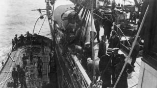 Groups of men on the deck of a ship which laying telegraph cable at sea, with the image showing men looking over the side of the ship at a smaller vessel carrying the cable, circa 1900