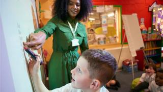 A black teacher helps a pupil write on a whiteboard
