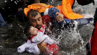 A Syrian refugee holds on to his children as he struggles to walk off a dinghy on the Greek island of Lesbos, 24 Aug