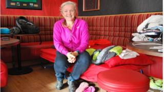 Gill Burns sitting on camp bed in Malone Rugby Club Bar area