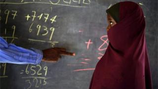 A young pupil stands next to her teacher as he points at mathematical sums on a blackboard