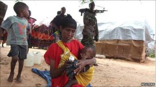 A displaced Somali woman sits in front of her makeshift shelter as a government soldier walks past in Madina district, southern Mogadishu, 3 August 2011