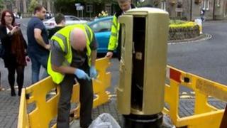 Crowd watches as a man paints a Royal Mail post box gold