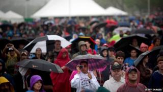 Hundreds of people stand with umbrellas and rain gear in front of a tent